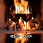 Two wine glasses stand side by side, on a table, in front of a fireplace.