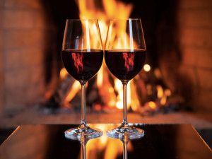 Two wine glasses stand side by side, on a table, in front of a fireplace.